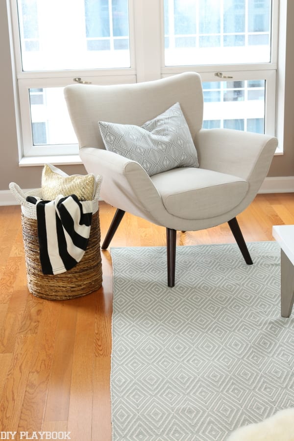 Welcoming greige armchair with a basket of throws and pillows include a black and white striped throw. 