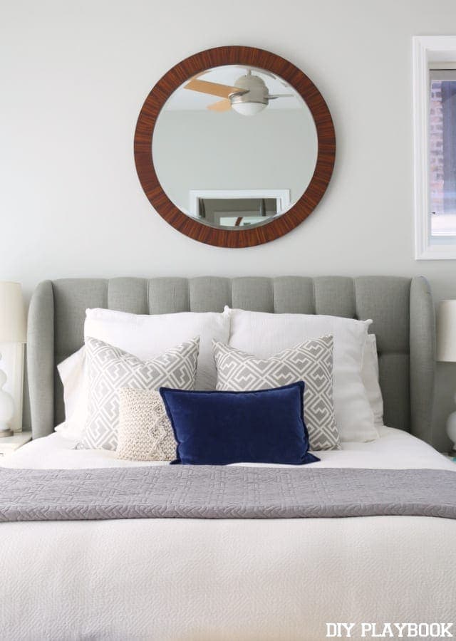 Bed with grey headboard and round mirror. 