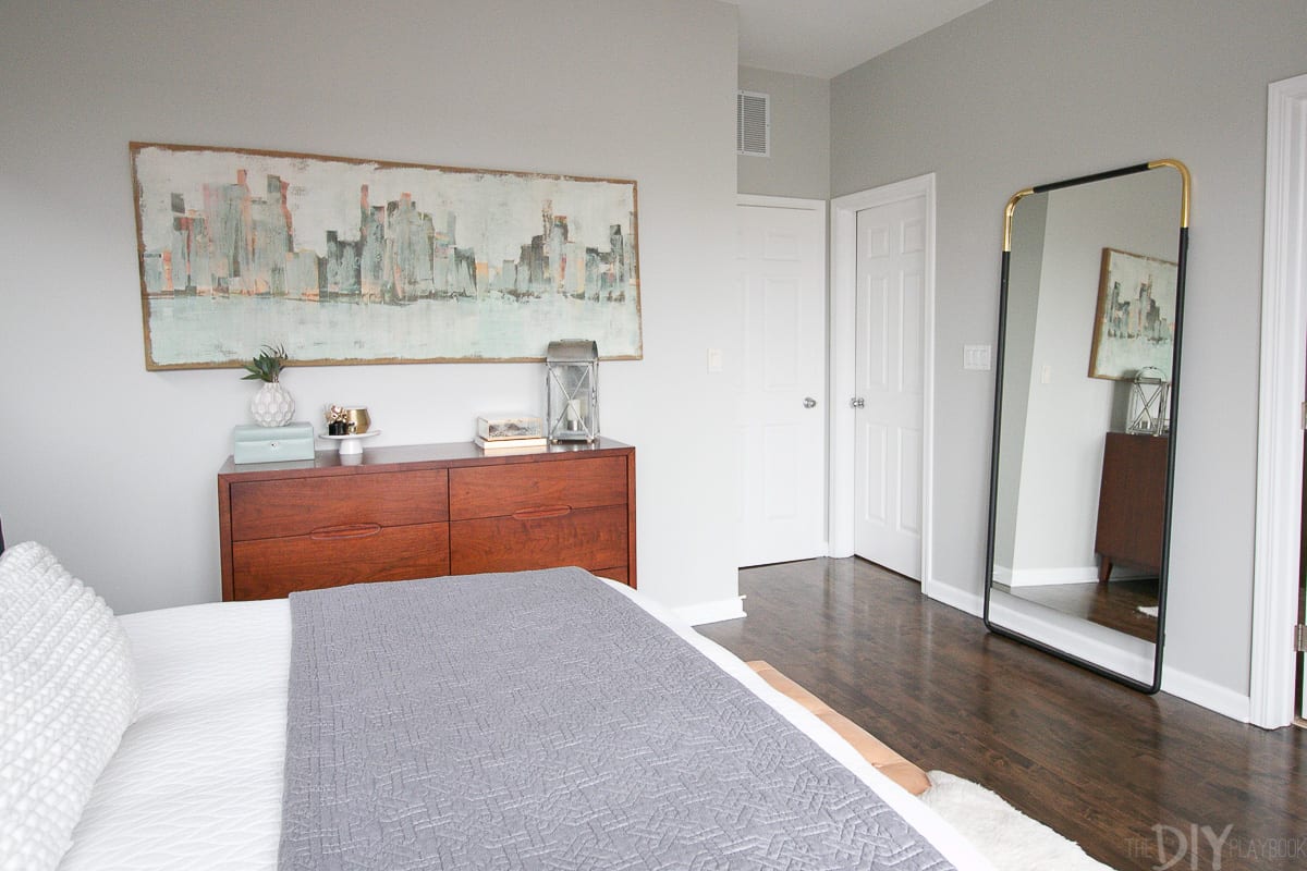 Light and airy master bedroom space with art, a full-length mirror, and a wood dresser.