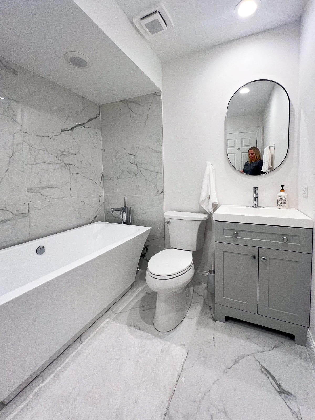Une salle de bain sans fenêtre avec sol en marbre, grande baignoire, carrelage en marbre aux murs et vanité grise